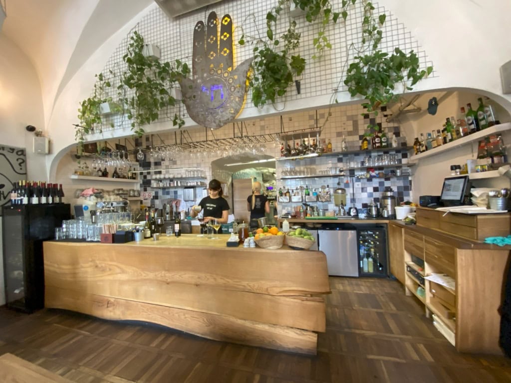 Innenraum eines Restaurants; im Zentrum eine Holzbar, dahinter bereitet eine Person in schwarzem Shirt ein Getränk zu. Über der Bar eine große Metallhand mit eingelassenem Auge, die »Hand der Fatima«