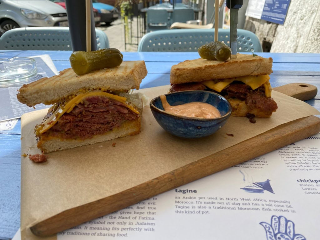 Auf einem Holzbrett liegen die zwei Hälften eines Pastrami-Sandwiches, darauf jeweils eine Gewürzgurke mit Holzpin befestigt. Zwischen den Hälften eine Schale mit Dip