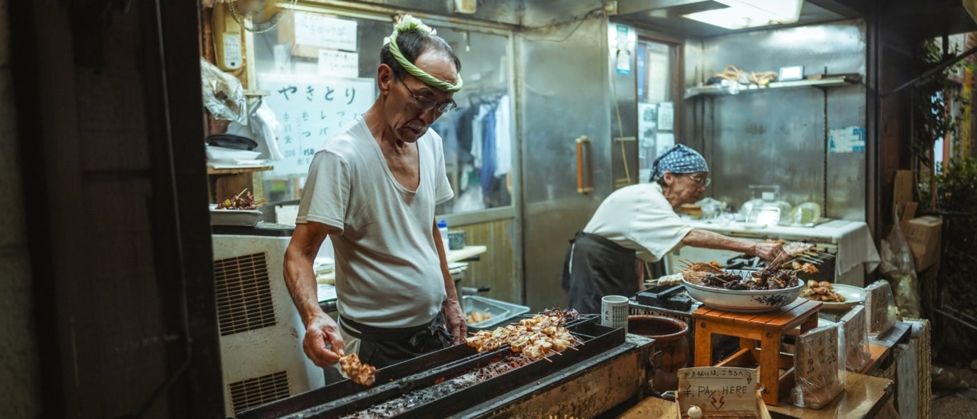 Ein älterer Mann und eine ältere Frau betreiben gemeinsam einen kleinen Imbissstand in Tokio und verkaufen Yakitori-Spieße.