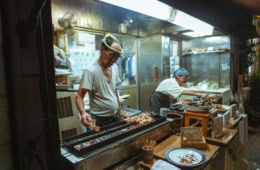 Ein älterer Mann und eine ältere Frau betreiben gemeinsam einen kleinen Imbissstand in Tokio und verkaufen Yakitori-Spieße.