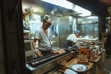 Ein älterer Mann und eine ältere Frau betreiben gemeinsam einen kleinen Imbissstand in Tokio und verkaufen Yakitori-Spieße.