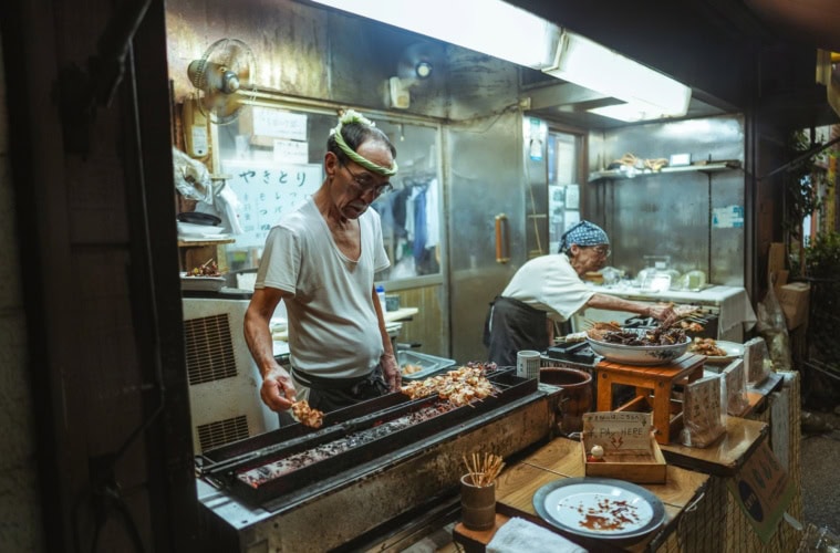 Ein älterer Mann und eine ältere Frau betreiben gemeinsam einen kleinen Imbissstand in Tokio und verkaufen Yakitori-Spieße.