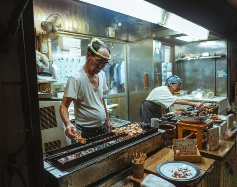 Ein älterer Mann und eine ältere Frau betreiben gemeinsam einen kleinen Imbissstand in Tokio und verkaufen Yakitori-Spieße.