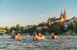 Eine Gruppe junger Menschen schwimmt an einem Sommertag im Rhein in Basel; im Hintergrund ragt der Dom aus den Dächern der Stadt hervor.