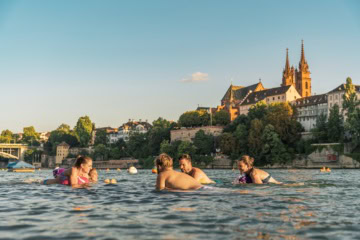 Eine Gruppe junger Menschen schwimmt an einem Sommertag im Rhein in Basel; im Hintergrund ragt der Dom aus den Dächern der Stadt hervor.