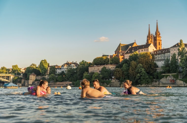 Eine Gruppe junger Menschen schwimmt an einem Sommertag im Rhein in Basel; im Hintergrund ragt der Dom aus den Dächern der Stadt hervor.