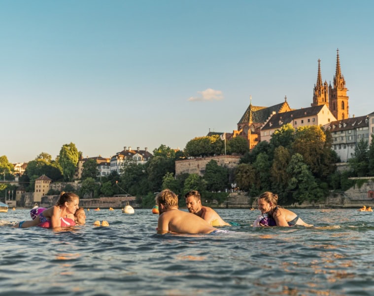 Eine Gruppe junger Menschen schwimmt an einem Sommertag im Rhein in Basel; im Hintergrund ragt der Dom aus den Dächern der Stadt hervor.