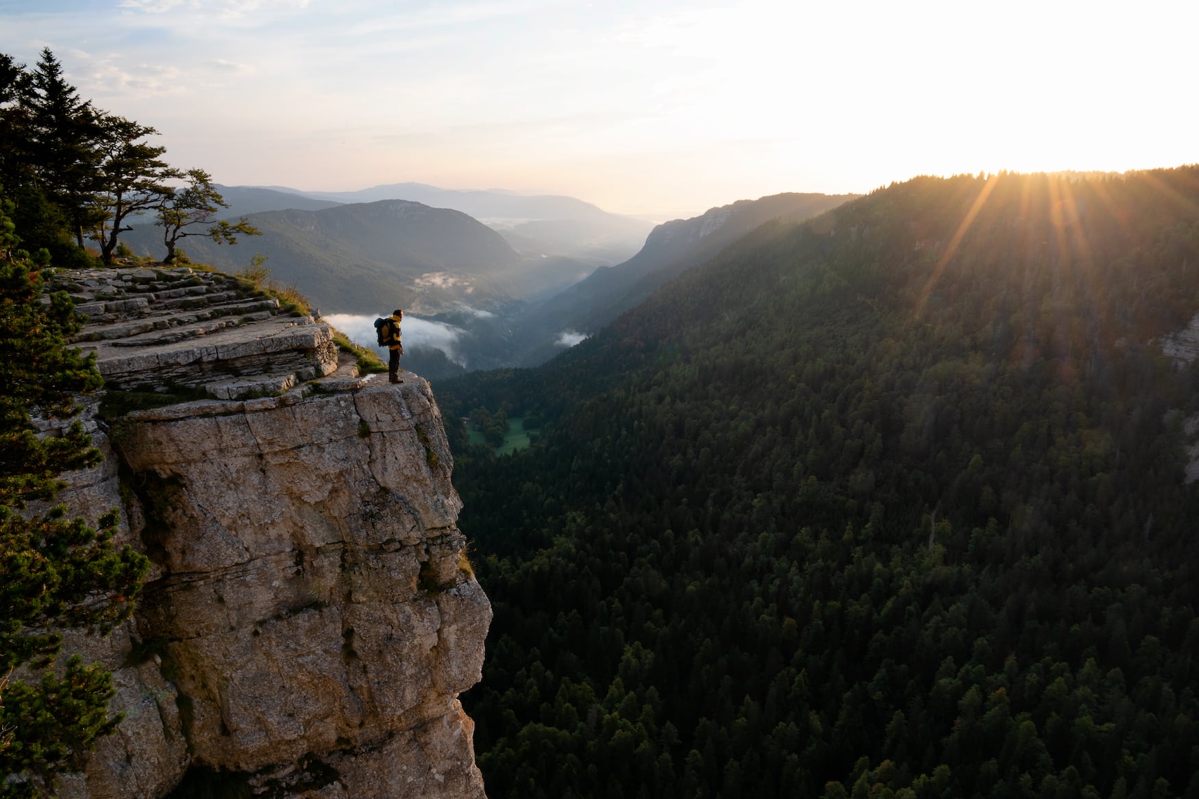 Sonnenaufgang am Creux du Van Neuchatel mit einem Mann, der auf einer steilen Klippe steht und dem warmen Sonnenlicht über dem Grat entgegensieht