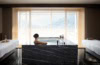 Eine Frau sitzt in einem Hotelspa in einem eckigen Whirpool mittig im Raum. Der Whirpool und der Boden sind aus schwarzem Marmor, links und rechts am Bildrand stehen zwei Liegen. Durch den weißen Vorhang vor dem mittigen Fenster lässt sich ein Alpenpanorama in der Sonne erkennen.