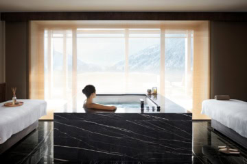Eine Frau sitzt in einem Hotelspa in einem eckigen Whirpool mittig im Raum. Der Whirpool und der Boden sind aus schwarzem Marmor, links und rechts am Bildrand stehen zwei Liegen. Durch den weißen Vorhang vor dem mittigen Fenster lässt sich ein Alpenpanorama in der Sonne erkennen.