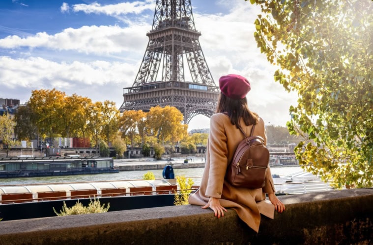 Eine Touristin mit Baskenmütze sitzt am Ufer der Seine und genießt die schöne Aussicht auf den Eiffelturm in Paris