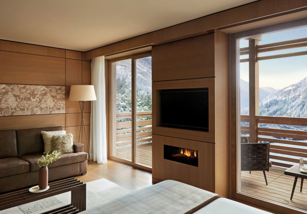 Ein Hotelzimmer mit holzverkleideten Wänden. Links im Bild eine braune Ledercouch, davor ein Metalltisch, unten am Bildrand die Ecke eines Bettes, rechts zwei bodentiefe Fenster mit einem Balkon mit Ausblick auf die Berge. In ein Wandstück zwischen den Fenstern ist ein Fernseher eingelassen, darunter ein möglicherweise künstlicher Kamin.