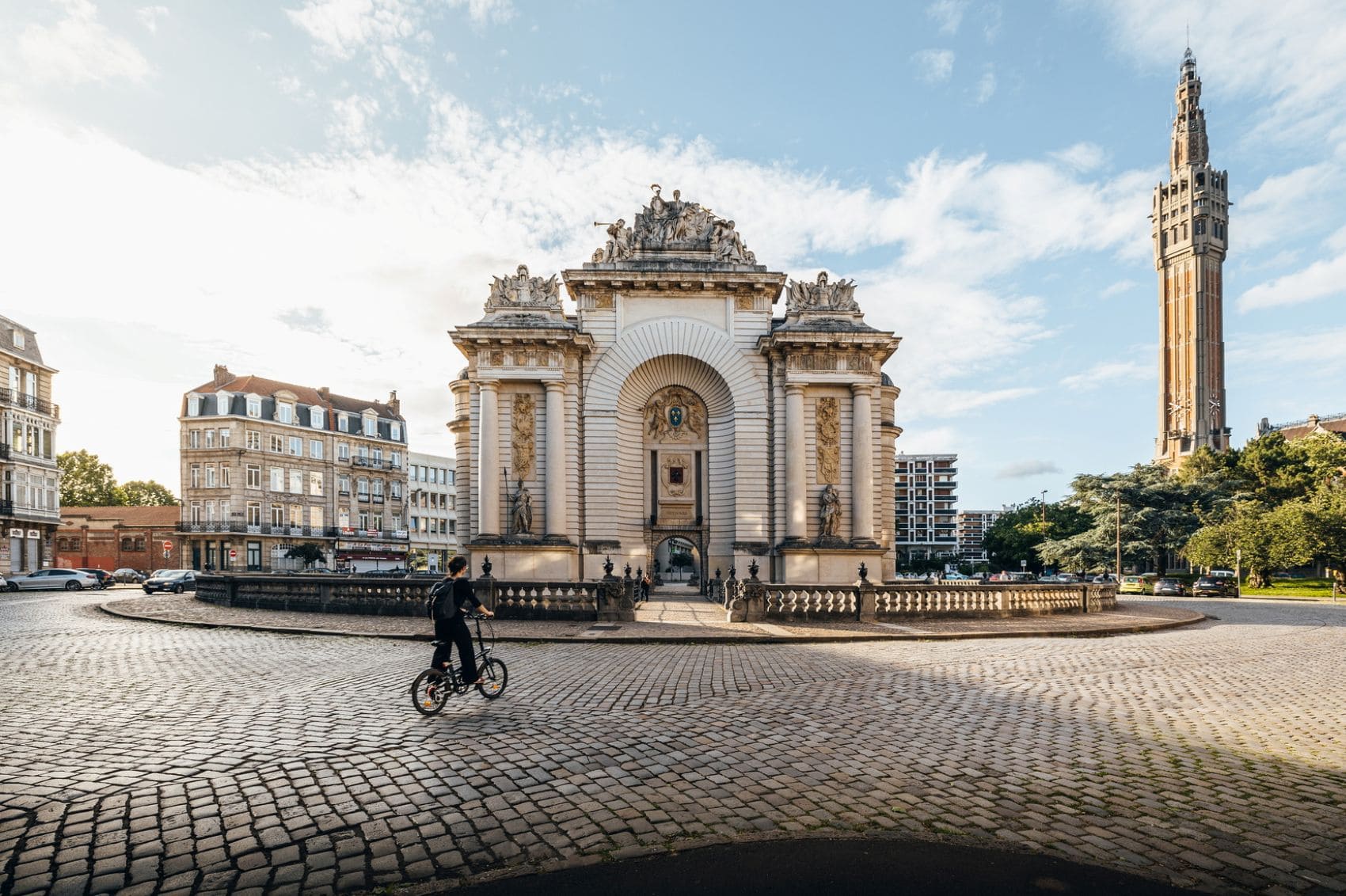 Junge fährt Fahrrad vor dem Porte de Paris in Lille, rechts ist der Belfried zu sehen