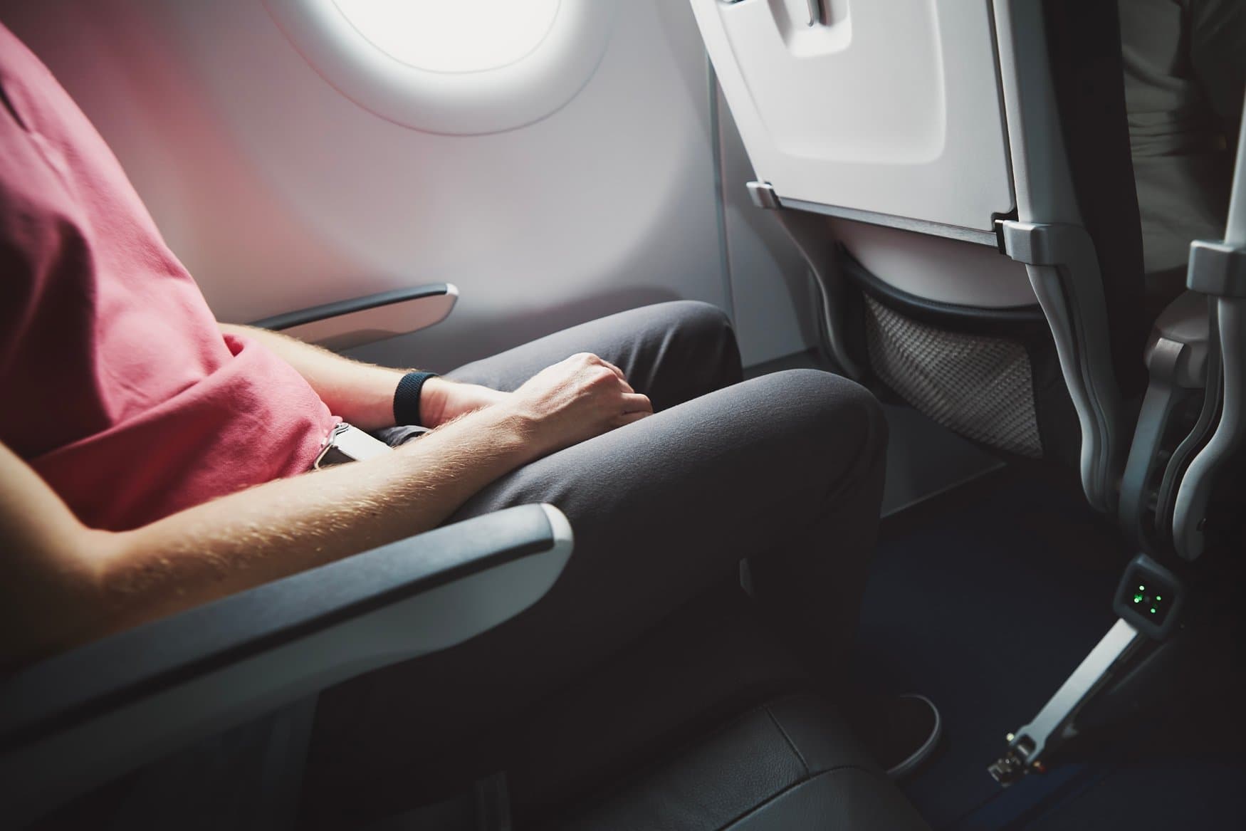 Ein Passagier in rotem Shirt sitzt am Fensterplatz eines Flugzeugs, die Arme auf den Armlehnen. Vor ihm ist der Abstand zur nächsten Sitzreihe mit hochgeklapptem Tabletttisch zu sehen.