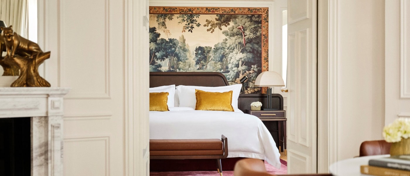 Zwei Altbauzimmer mit Dielenboden und hohen Decken, die über eine weite Doppeltür verbunden sind. Der Blick geht aus einem Zimmer ins andere. Im Zimmer des Betrachters steht zur rechten ein niedriger Holzstuhl mit abgerundeter Polsterlehne, zur Linken ist der Rand eines Marmorkamins mit Spiegel darüber zu sehen. Im zweiten Zimmer steht ein breites Doppelbett, dahinter ein wandhohes Renaissancegemälde eines Waldes. Das Bettzeug is strahlend weiß, mit Ausnahme zweier gold-gelber Samtzierkissen. Alle Wände sind weiß gestrichen, die Zimmer wirken sehr hell.