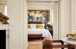 Zwei Altbauzimmer mit Dielenboden und hohen Decken, die über eine weite Doppeltür verbunden sind. Der Blick geht aus einem Zimmer ins andere. Im Zimmer des Betrachters steht zur rechten ein niedriger Holzstuhl mit abgerundeter Polsterlehne, zur Linken ist der Rand eines Marmorkamins mit Spiegel darüber zu sehen. Im zweiten Zimmer steht ein breites Doppelbett, dahinter ein wandhohes Renaissancegemälde eines Waldes. Das Bettzeug is strahlend weiß, mit Ausnahme zweier gold-gelber Samtzierkissen. Alle Wände sind weiß gestrichen, die Zimmer wirken sehr hell.