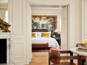 Zwei Altbauzimmer mit Dielenboden und hohen Decken, die über eine weite Doppeltür verbunden sind. Der Blick geht aus einem Zimmer ins andere. Im Zimmer des Betrachters steht zur rechten ein niedriger Holzstuhl mit abgerundeter Polsterlehne, zur Linken ist der Rand eines Marmorkamins mit Spiegel darüber zu sehen. Im zweiten Zimmer steht ein breites Doppelbett, dahinter ein wandhohes Renaissancegemälde eines Waldes. Das Bettzeug is strahlend weiß, mit Ausnahme zweier gold-gelber Samtzierkissen. Alle Wände sind weiß gestrichen, die Zimmer wirken sehr hell.