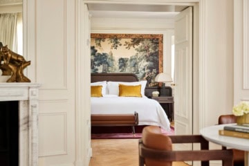 Zwei Altbauzimmer mit Dielenboden und hohen Decken, die über eine weite Doppeltür verbunden sind. Der Blick geht aus einem Zimmer ins andere. Im Zimmer des Betrachters steht zur rechten ein niedriger Holzstuhl mit abgerundeter Polsterlehne, zur Linken ist der Rand eines Marmorkamins mit Spiegel darüber zu sehen. Im zweiten Zimmer steht ein breites Doppelbett, dahinter ein wandhohes Renaissancegemälde eines Waldes. Das Bettzeug is strahlend weiß, mit Ausnahme zweier gold-gelber Samtzierkissen. Alle Wände sind weiß gestrichen, die Zimmer wirken sehr hell.