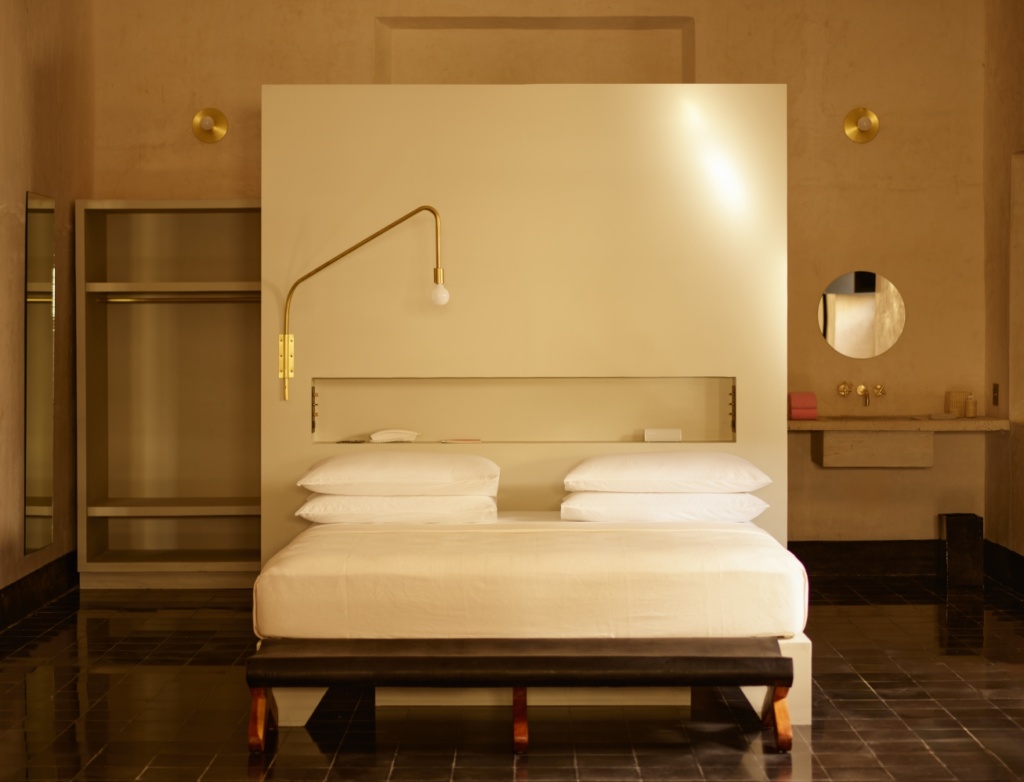 In einem Raum steht mittig ein flaches Holzbett mit weißer Matratze und weißen Bezügen auf einem schwarzen Fliesenboden. Hinter dem Bett ein hoher Raumtrenner aus blankem Beton, dahinter links ein offener, grauer Schrank und rechts ein Waschbecken aus Beton.