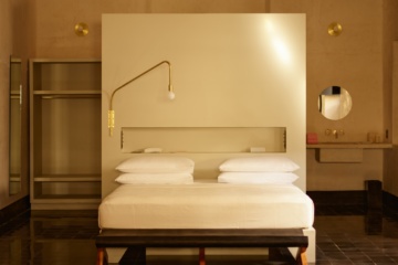 In einem Raum steht mittig ein flaches Holzbett mit weißer Matratze und weißen Bezügen auf einem schwarzen Fliesenboden. Hinter dem Bett ein hoher Raumtrenner aus blankem Beton, dahinter links ein offener, grauer Schrank und rechts ein Waschbecken aus Beton.