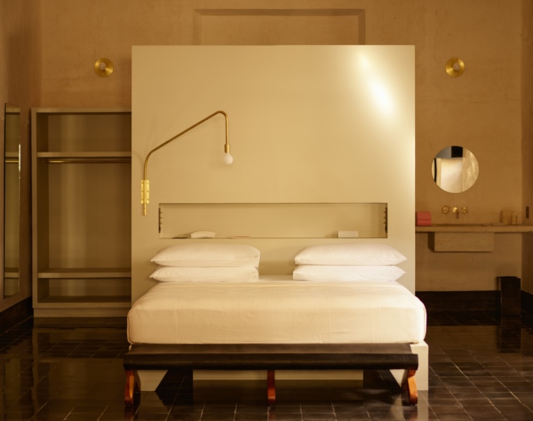 In einem Raum steht mittig ein flaches Holzbett mit weißer Matratze und weißen Bezügen auf einem schwarzen Fliesenboden. Hinter dem Bett ein hoher Raumtrenner aus blankem Beton, dahinter links ein offener, grauer Schrank und rechts ein Waschbecken aus Beton.