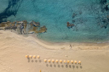 Luftaufnahme eines hellen Sandstrands mit türkisfarbenem, klarem Wasser und Felsen im Meer. Mehrere Sonnenschirme stehen in einer Reihe am Strand, vereinzelt liegen Menschen im Sand oder schwimmen im Wasser