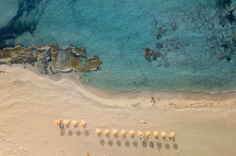 Luftaufnahme eines hellen Sandstrands mit türkisfarbenem, klarem Wasser und Felsen im Meer. Mehrere Sonnenschirme stehen in einer Reihe am Strand, vereinzelt liegen Menschen im Sand oder schwimmen im Wasser