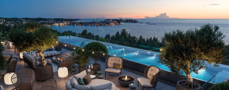 Blick auf die Rooftop-Bar eines Strandhotels am Abend. Zur Linken die Terrasse mit mehreren Sitzgruppen, Polster- und Bastmöbel gemischt. Rechts dahinter ein langezogener Infinitypool. Am Horizont ist die Stadt Porec zu sehen, dahinter das blaue Mittelmeer.