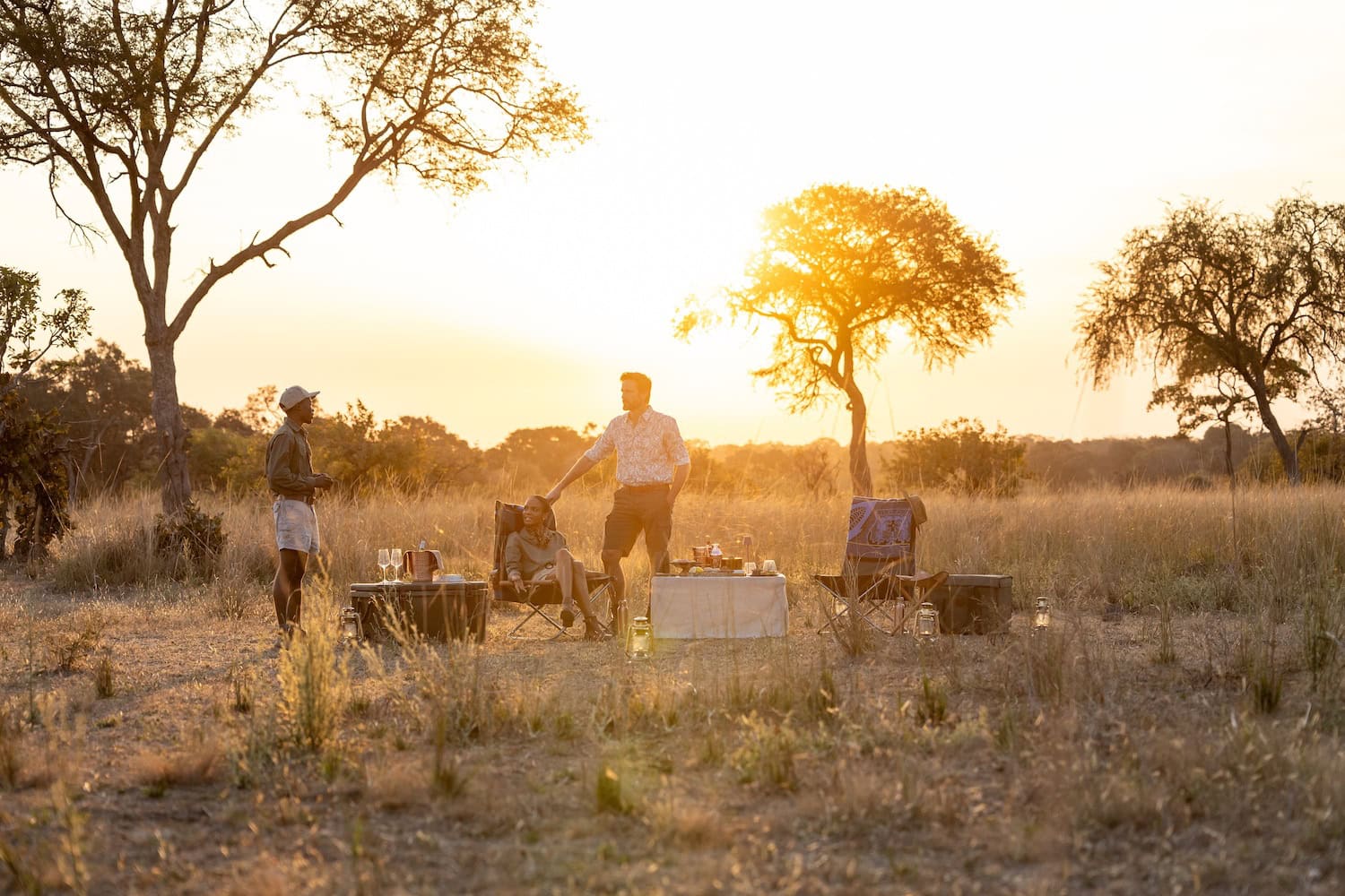 Anantara Kafue River Paar in der Abendsonne beim Picknick in der Wildnis in Sambia im Gespräch mit einem Ranger, hohes Gras, einzelne Bäume
