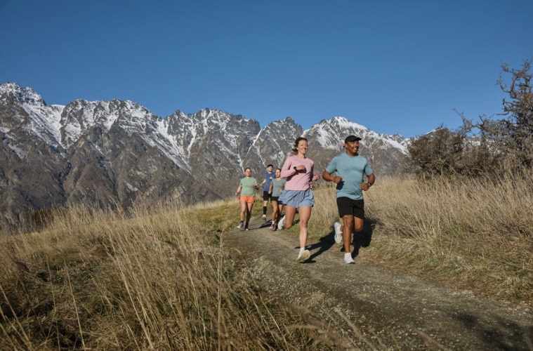 Zwei Läufer auf einem Naturtrail zwischen hohen Gräsnern, im Hintergrund hohe, schneebedeckte Berge in Neuseeland