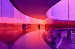 Eine Person geht durch einen kreisrunden Skywalk mit rosa-lilafarbenen Glaswänden über den Dächern von Aarhus