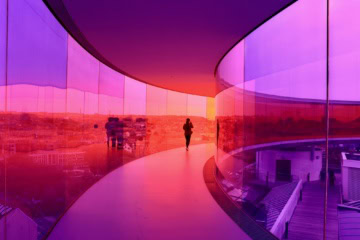 Eine Person geht durch einen kreisrunden Skywalk mit rosa-lilafarbenen Glaswänden über den Dächern von Aarhus
