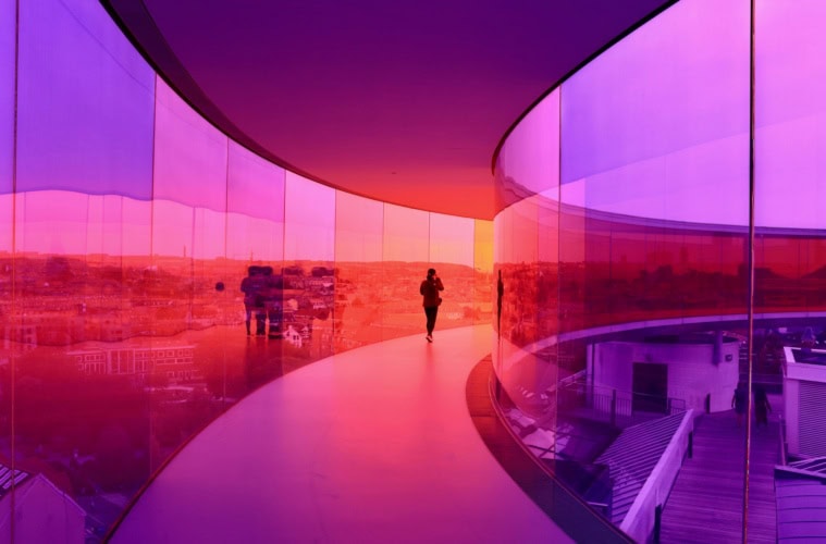 Eine Person geht durch einen kreisrunden Skywalk mit rosa-lilafarbenen Glaswänden über den Dächern von Aarhus