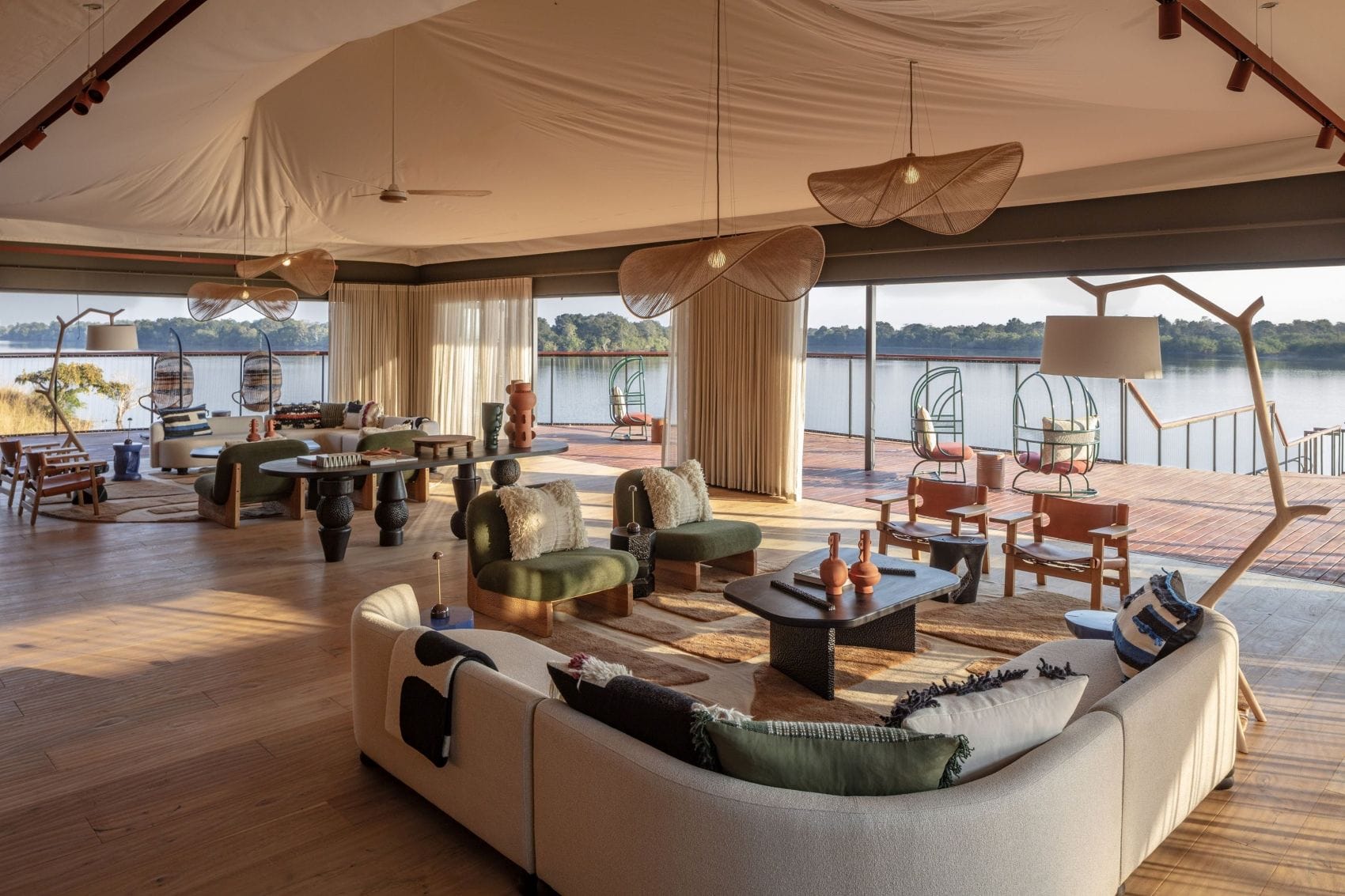 Großzügige Lounge im Anantara Tented Camp Kafue River in Sambia mit geschwungenen Sofas, Flechtleuchten, Holzboden und Panoramablick auf den Fluss.