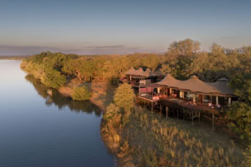 Luftaufnahme des Anantara Tented Camp Kafue River in Sambia mit luxuriösen Zeltvillen auf Stelzen, Terrassen und bewaldetem Flussufer im Abendlicht.