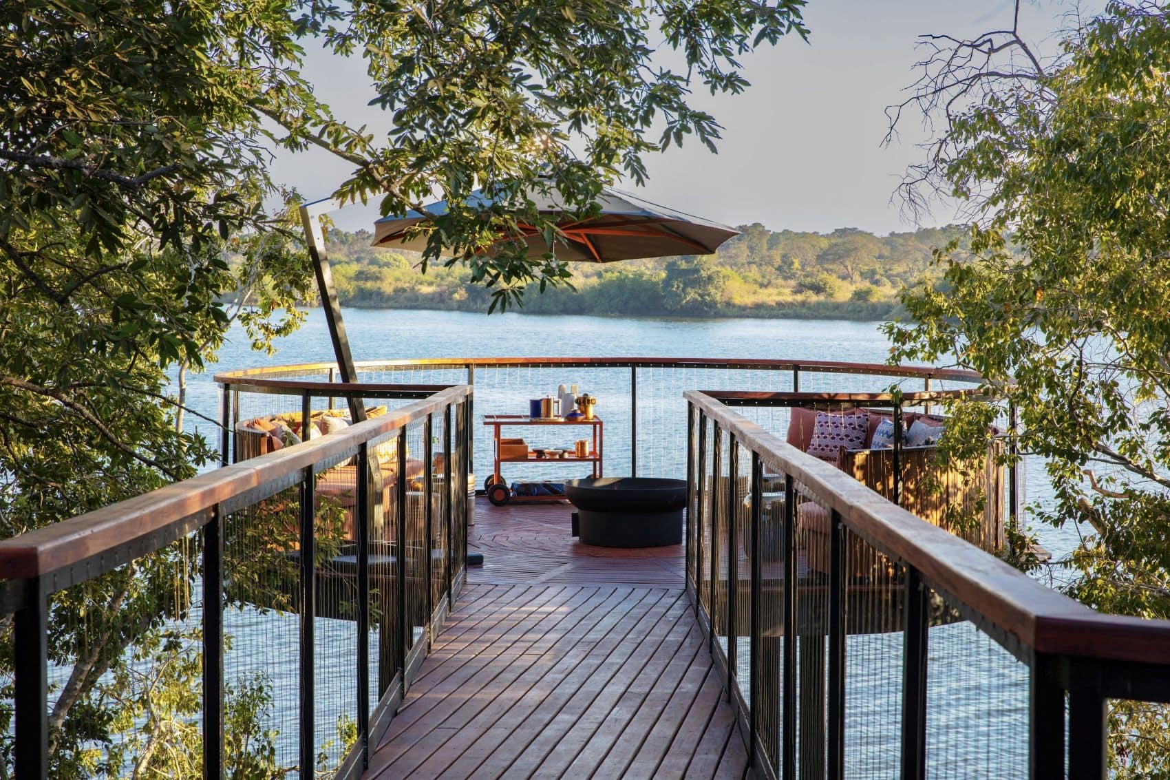 Holzsteg zu einer erhöhten Terrasse mit Sitzgelegenheiten, Sonnenschirm und Blick auf den Kafue-Fluss im Anantara Tented Camp in Sambia.
