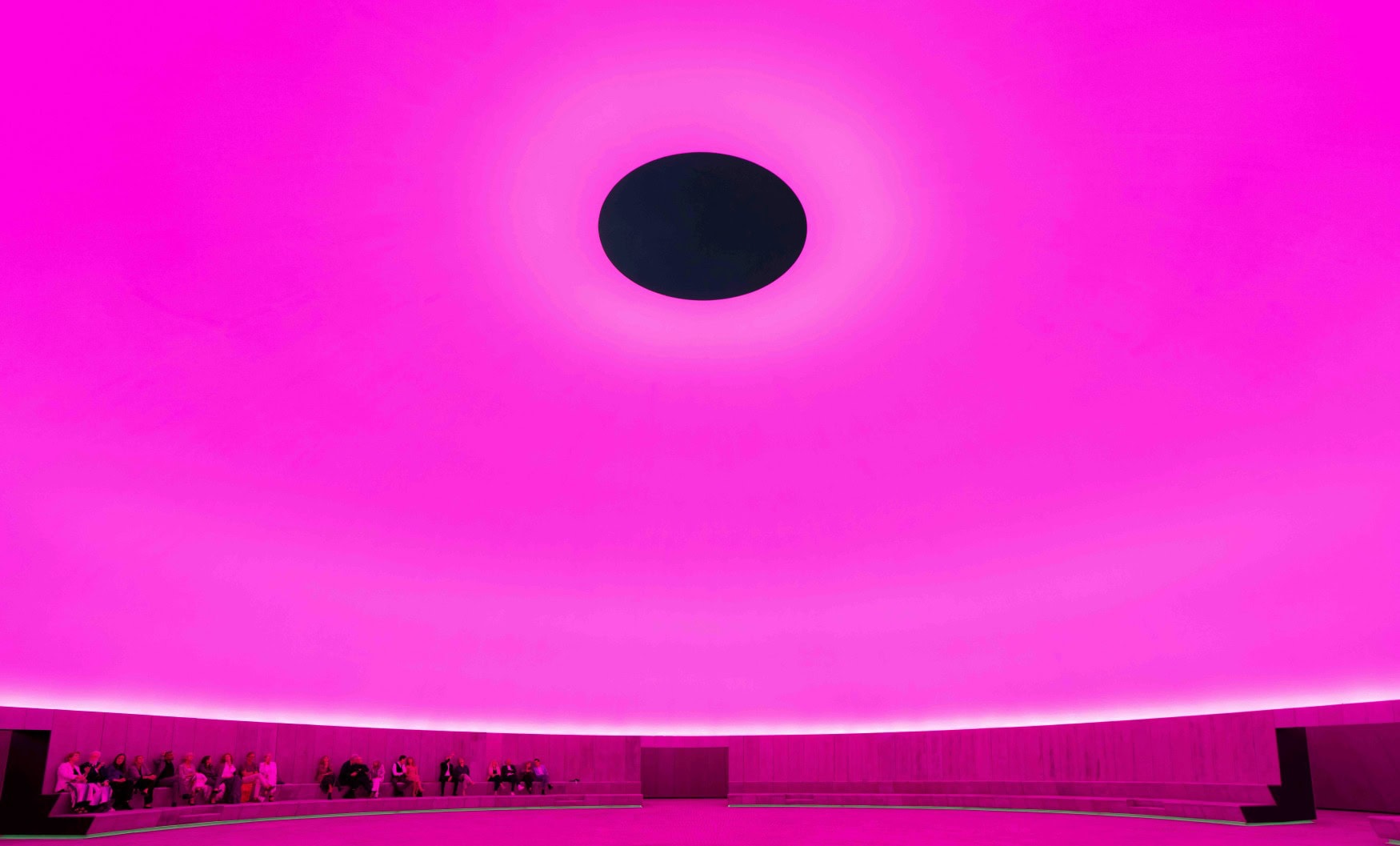 Die Kunstinstallation "As Seen Below" des Künstlers James Turrell im Museum Aros in Aarhus; unter einer nahtlos scheinenden Kuppel, die in dunklem Lila angestrahlt wird, sitzen einige Menschen und blicken nach oben. Genau in der Mitte der Kuppel prangt ein runder, schwarzer Kreis