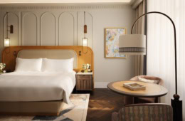 Zimmer in einem Luxushotel; zur Linken ein breites Doppelbett mit breitem, hölzernen Kopfteil auf einem grauen Teppich. Der Boden ist mit dunklem Parkett verkleidet. Zur rechten vor einem Fenster zwei elegante Polstersessel an einem runden Beistelltisch, darüber eine geschwungene Stehlampe. Zur rechten kommt durch ein Fenster hinter einem weißen Vorhang Tageslicht herein.