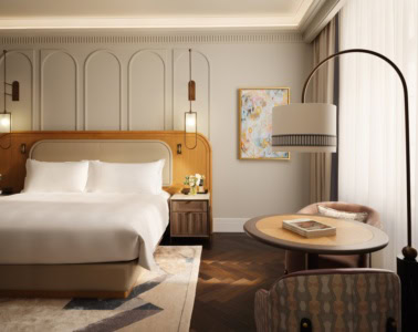 Zimmer in einem Luxushotel; zur Linken ein breites Doppelbett mit breitem, hölzernen Kopfteil auf einem grauen Teppich. Der Boden ist mit dunklem Parkett verkleidet. Zur rechten vor einem Fenster zwei elegante Polstersessel an einem runden Beistelltisch, darüber eine geschwungene Stehlampe. Zur rechten kommt durch ein Fenster hinter einem weißen Vorhang Tageslicht herein.