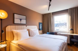 Doppelbett mit weißen Bezügen in einem Hotelzimmer; die Wand hinter dem Bett ist hellbraun gestrichen, zwei kleine Bilder hängen daran. Rechts vom Bett lässt ein großes Fenster Licht herein. Am Fenster steht eine schmaler Schreibtisch, links und rechts daneben zwei blaue Polsterstühle.