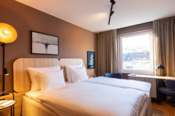 Doppelbett mit weißen Bezügen in einem Hotelzimmer; die Wand hinter dem Bett ist hellbraun gestrichen, zwei kleine Bilder hängen daran. Rechts vom Bett lässt ein großes Fenster Licht herein. Am Fenster steht eine schmaler Schreibtisch, links und rechts daneben zwei blaue Polsterstühle.