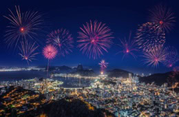 Luftaufnahme von Rio de Janeiro bei Nacht; über der hell erleuchteten Statt mit dem Zuckerhut im Hintergrund steigt ein riesiges Feuerwerk auf.