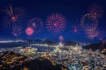 Luftaufnahme von Rio de Janeiro bei Nacht; über der hell erleuchteten Statt mit dem Zuckerhut im Hintergrund steigt ein riesiges Feuerwerk auf.