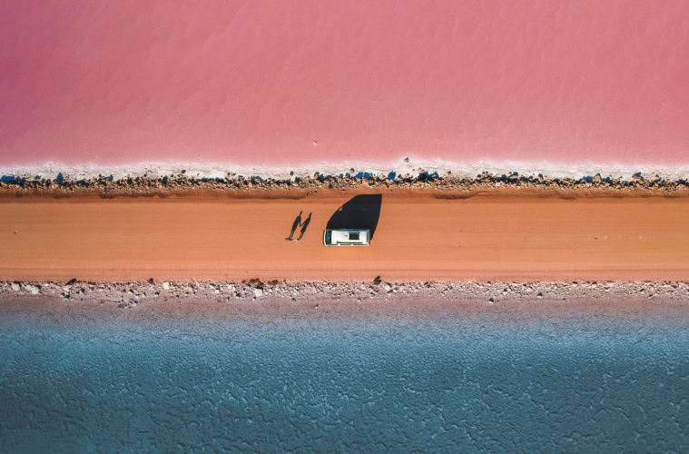 Luftaufnahme einer Straße, darauf ein Auto und zwei Personen. Nördlich der Straße ist ein rosa See, südlich ein blauer See