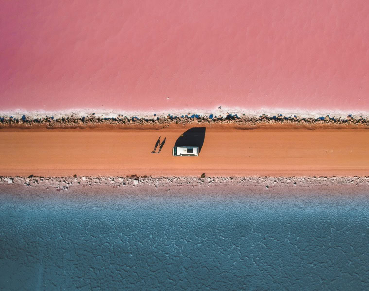 Luftaufnahme einer Straße, darauf ein Auto und zwei Personen. Nördlich der Straße ist ein rosa See, südlich ein blauer See