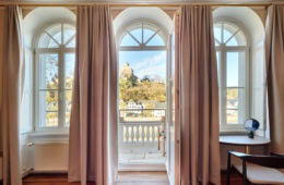 Zwei hohe Fenster und eine mittige Tür trennen ein Altbauzimmer mit Parkettboden von einem weoßen Balkon. Der Blick geht auf eine Burgruine in einem Wald.