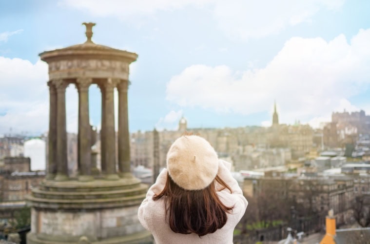 Frau von hinten mit dunklen Haaren, weißer Baskenmütze und weissem Mantel fotografiert in Edinburgh die Stadt von einem Hügel aus. Im Vordergrund das Dugald Stewart Monument auf dem Carlton Hill.