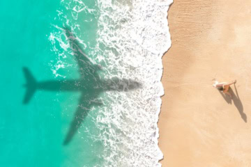 Schatten eines Flugzeugs über karibisch blauem Meer am Strand, Foto aus Vogelperspektive mit einer einzelnen Frau, die im Sand sitzt.