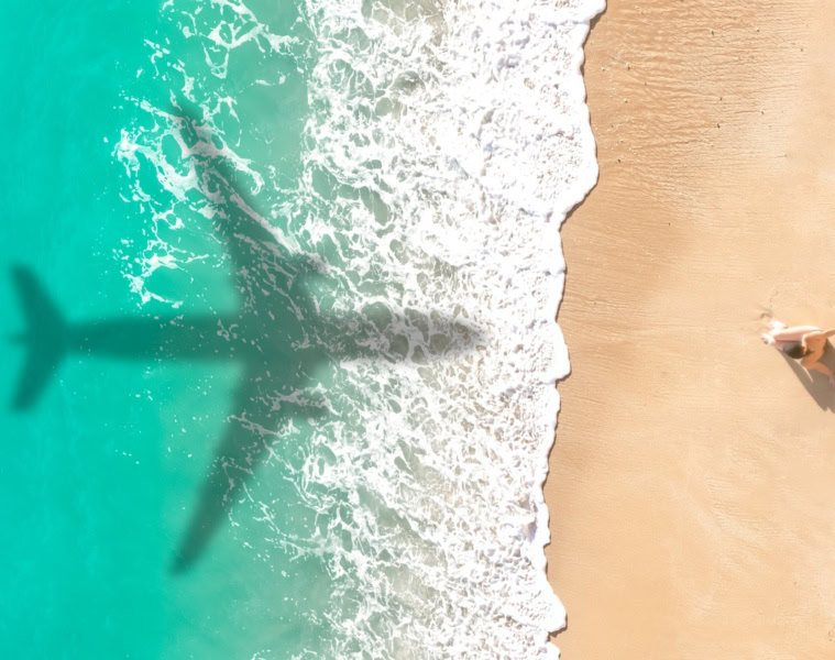 Schatten eines Flugzeugs über karibisch blauem Meer am Strand, Foto aus Vogelperspektive mit einer einzelnen Frau, die im Sand sitzt.