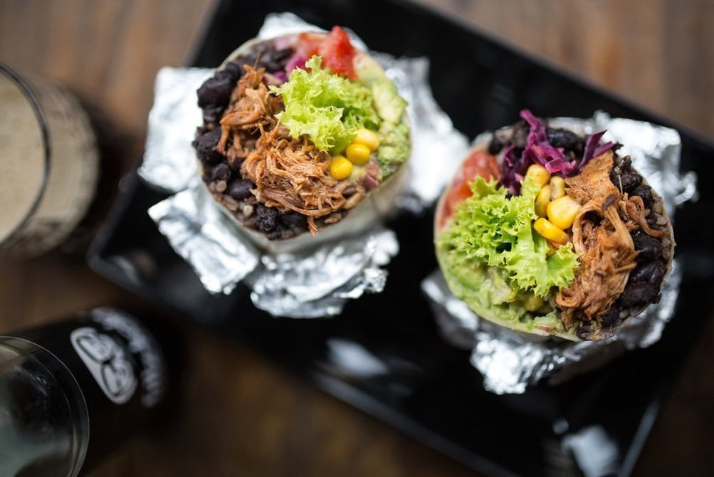 Aufsicht auf zwei aufgeschnittene Burritos in Alufolie auf einem schwarzen Serviertablett. In der Füllung sind Pulled Pork, schwarze Bohnen, Mais, Salat, Rotkohl und Guacamole zu sehen, links steht ein Glas dunkles Bier.